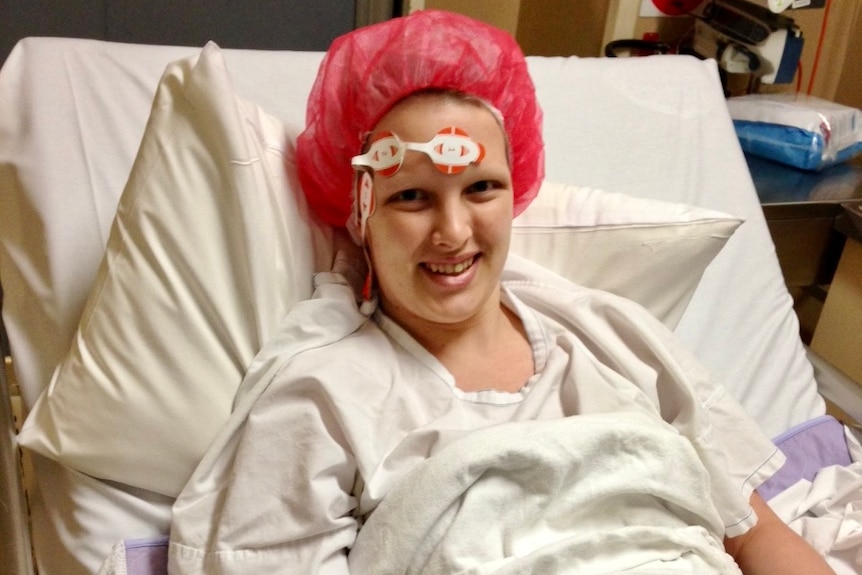 Kathryn woods in a hospital bed wearing a white gown and red hair net, with a machine connected to her.