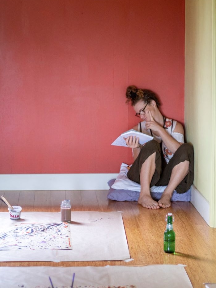 A woman, barefoot, sits in a corner reading a notebook. There are painting materials and paper on the floor nearby.