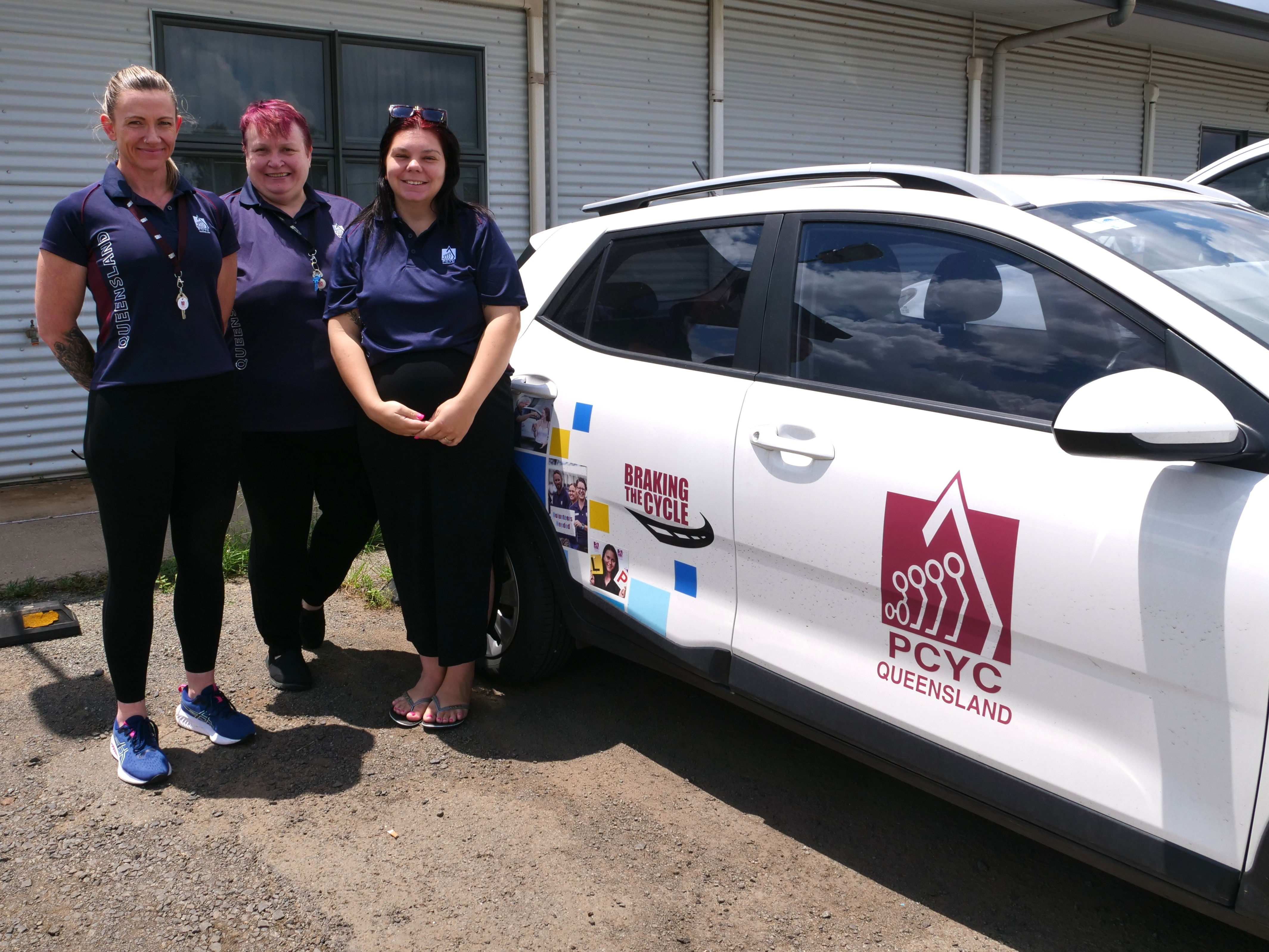 Três mulheres vestindo camisetas da marca PCYC, em frente a um pequeno carro com placas, em frente ao prédio da academia 