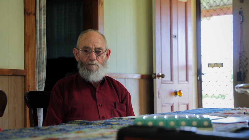 A balding elderly man with short grey hair and a  beard sits at a dining room table in a red button-up shirt and glasses.