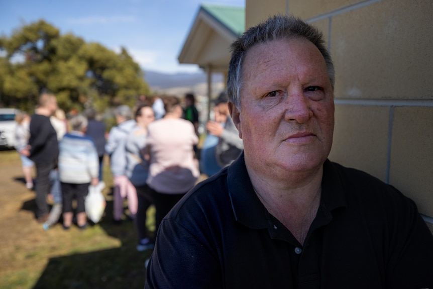 A man leans against a wall as a crowd of people mill around behind him.