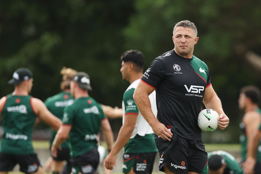 An NRL assistant coach looks over a field carrying a ball in one hand as players stand behind him at a distance.