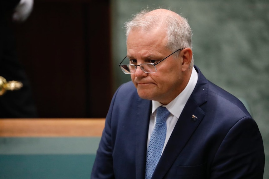 A man in a suit with grey hair and glasses looks solemn.