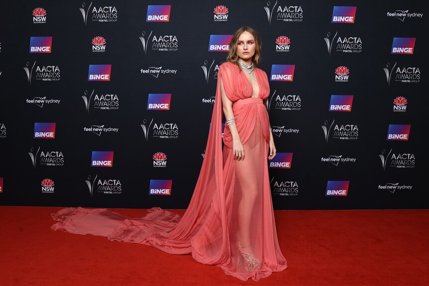 Olivia DeJonge, a white woman with mousey brown hair, wears a flowing coral-coloured dress on a red carpet.