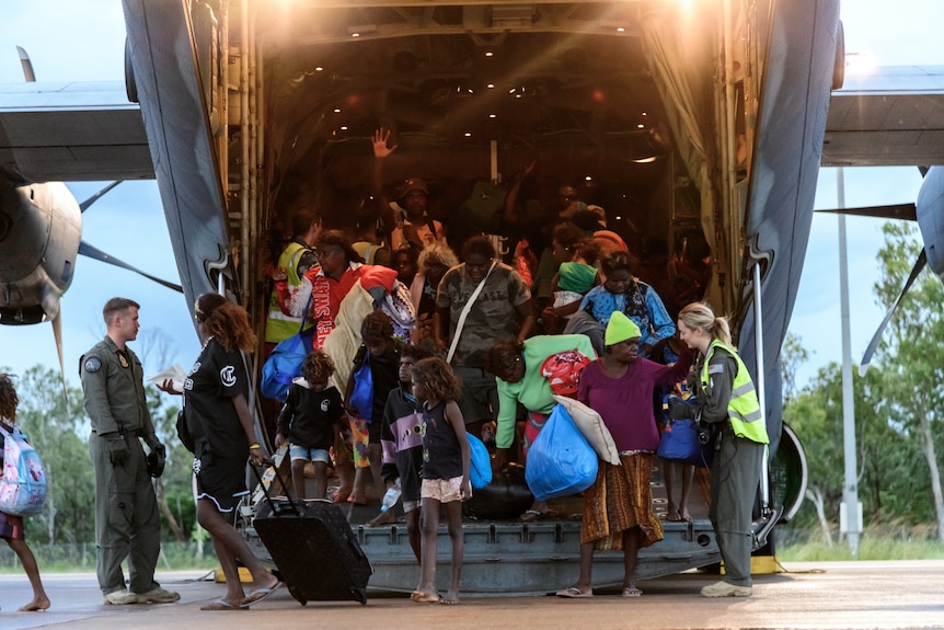 A group of people getting off a large plane, carrying bags and other items, and escorted by several army representives.