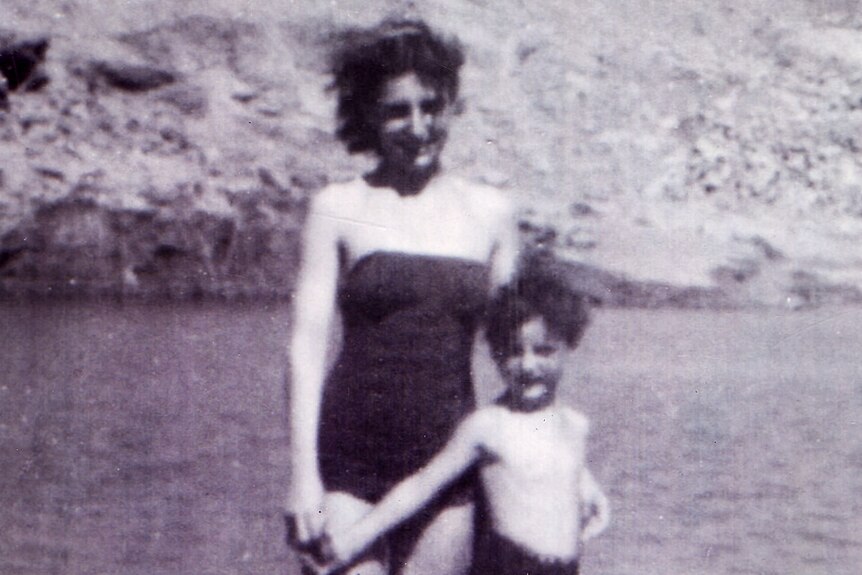 Black and white photo of young Rai next to his mother in front of the water at the beach, smiling, wearing swimming costumes.