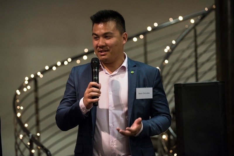 Sean smiles while speaking into a microphone at an event, wearing a suit.