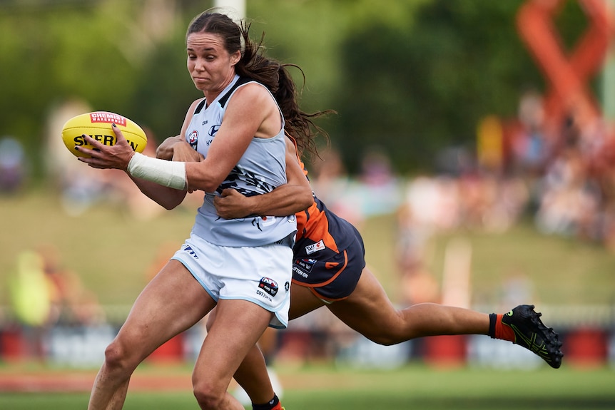An AFLW player grimaces as she holds the football while being tackled around the waist by a defender.