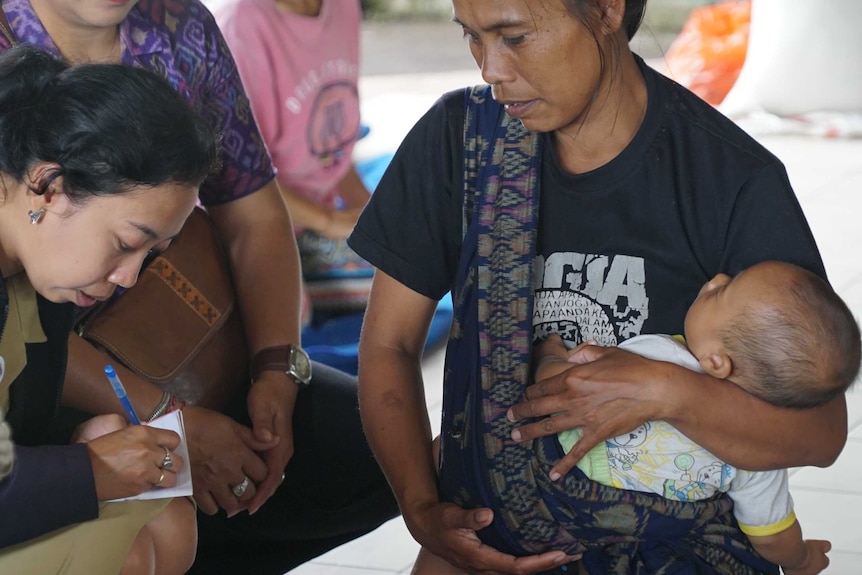 A midwife, sitting with a woman holding a baby, writes on a notebook.