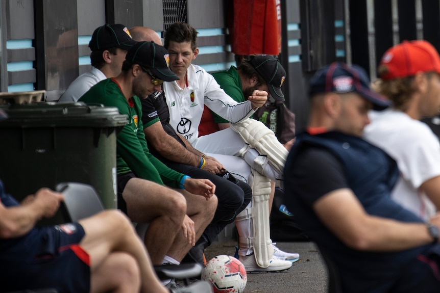 A cricketer wearing pads turns to chat to a man next to him as he waits to go into bat during a game.