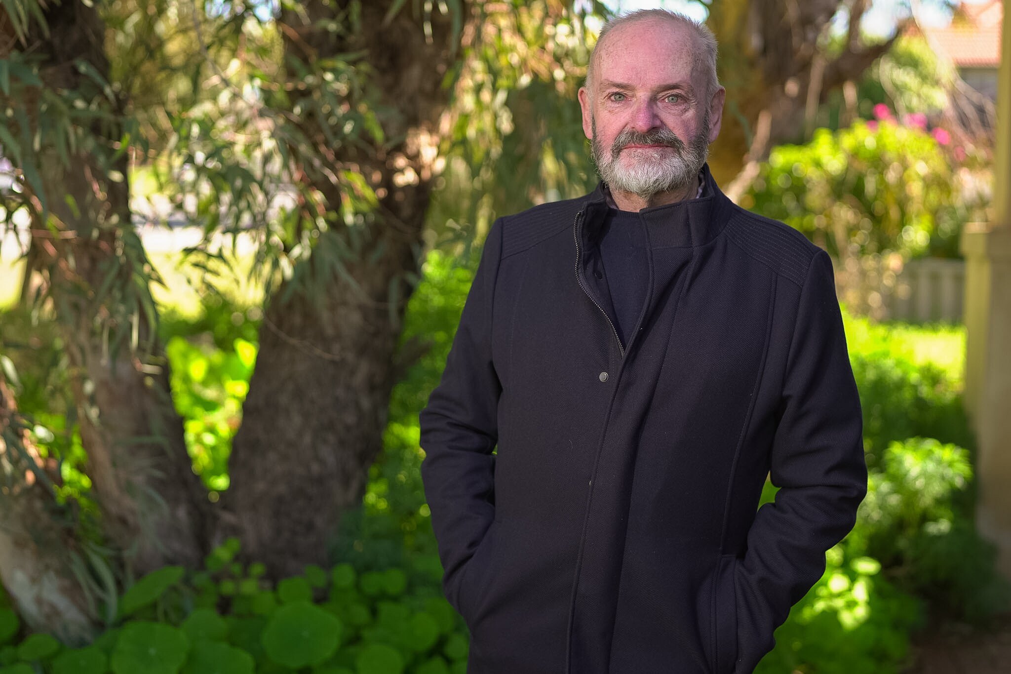Un hombre se para con un abrigo negro contra un jardín verde 