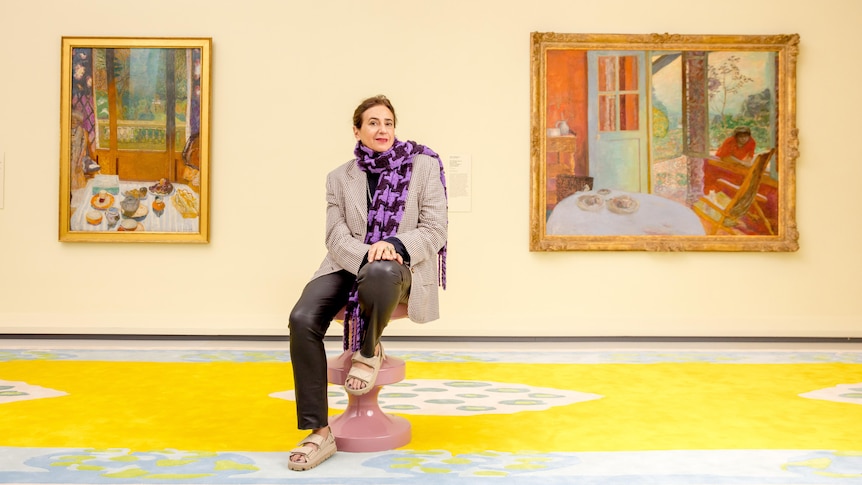 A woman with dark hair sits on a stool in front of two paintings, in a gallery room with a yellow floor.