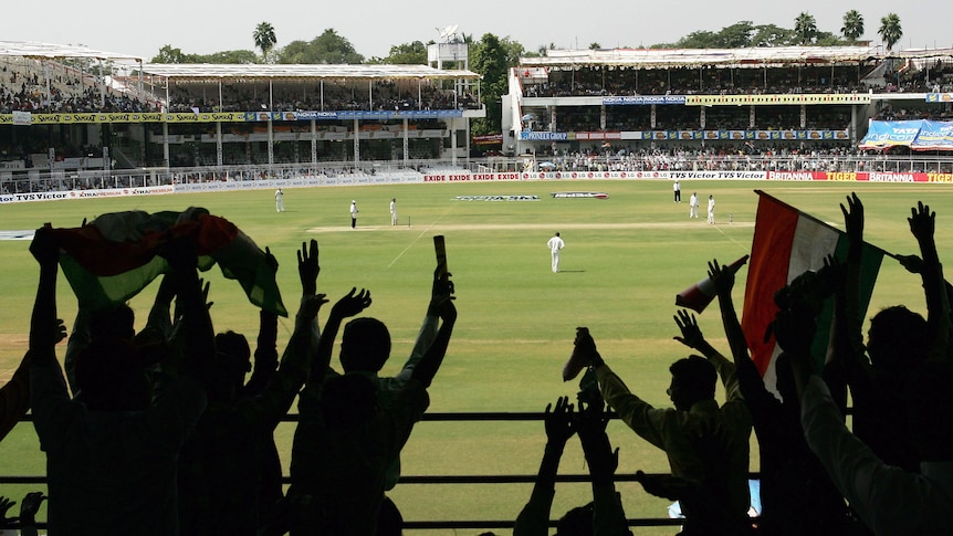 Fans cheer while looking out at the cricket pitch as seen from behind