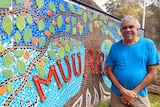 A man in a blue T-shirt stands next to a wall at the front of a building which features a colourful mosaic and says 'Muurrbay'.