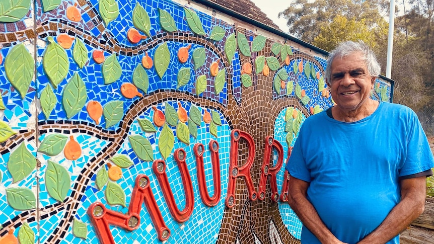 A man in a blue T-shirt stands next to a wall at the front of a building which features a colourful mosaic and says 'Muurrbay'.