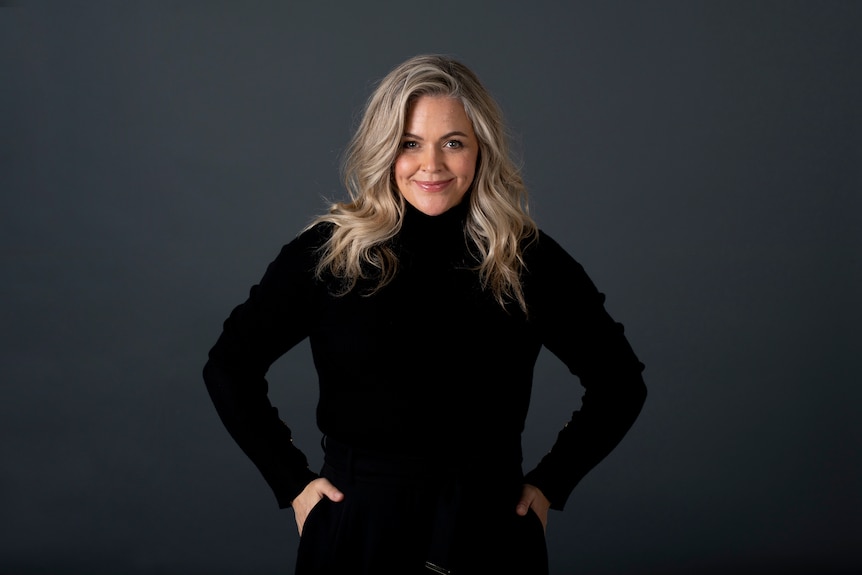 A professional headshot of Taryn Brumfitt smiling to the camera. She wears a black turtleneck jumper and black pants.