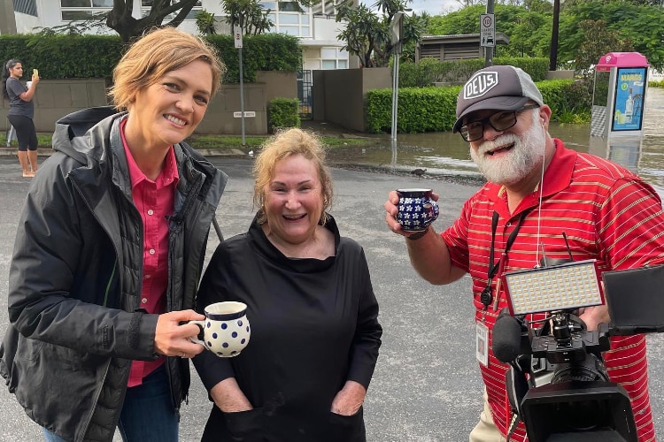 Journalist and cameraman holding a cup of tea each with a woman in between them.