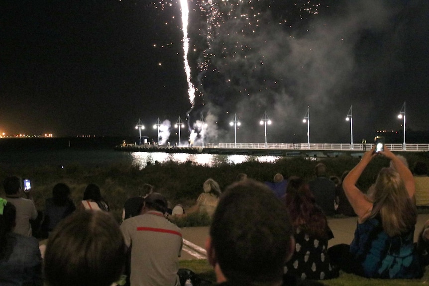 People sit on the grass as fireworks go off over a jetty.