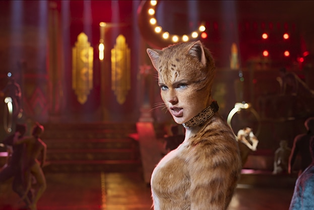 An fawn coloured CGI cat with woman's face stands in a dimly lit 1930s style cabaret-style music hall full of other cats.