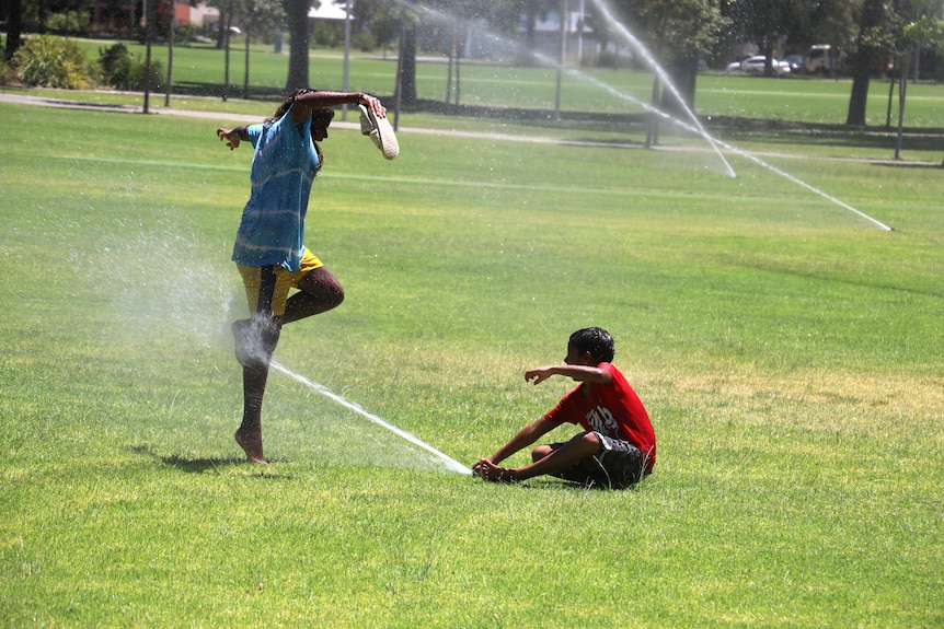 Two Indigenous children play under a sprinkler in a park, one seated and one standing with arms outstretched