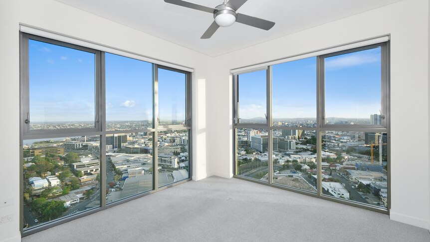 The corner of an empty room, grey carpet, white walls and windows with views of a city