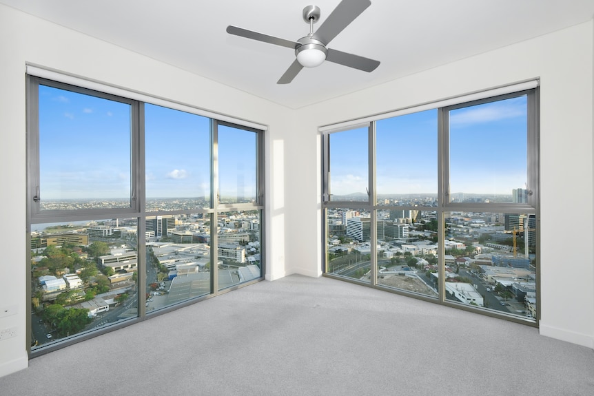 The corner of an empty room, grey carpet, white walls and windows with views of a city