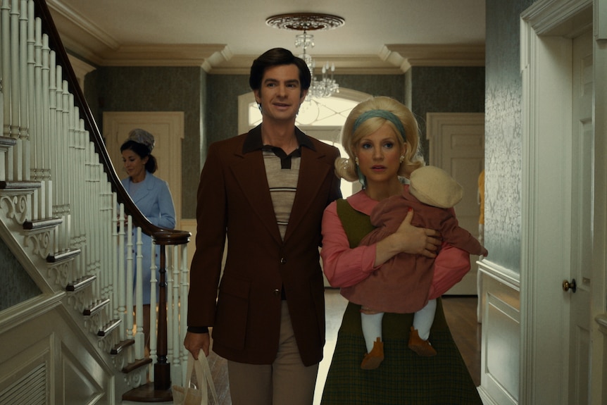 Middle-aged man and woman dressed in 50s attire walk through elegantly decorated house foyer. Woman carries baby in arms.