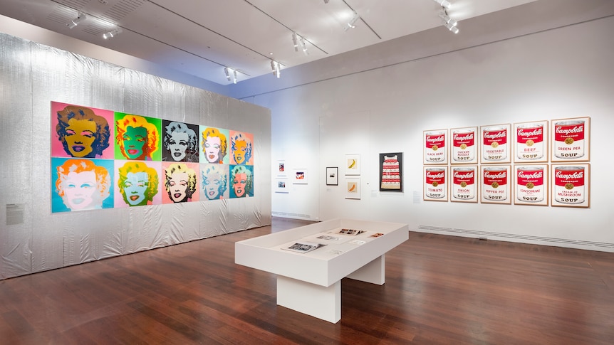 A gallery space, on the left are 10 pop art portraits of marilyn monroe in front of a silver wall, right campbell soup cans