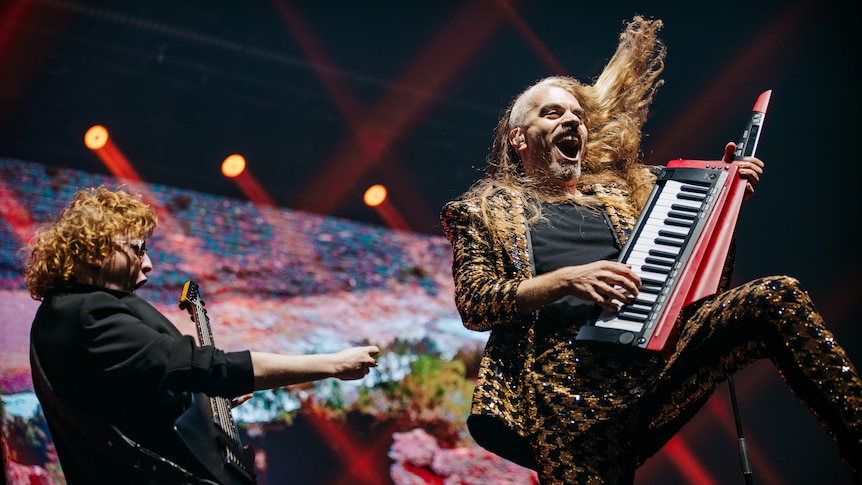 Two musicians on stage, one playing a guitar and the other a keytar, during a performance of a song in concert.