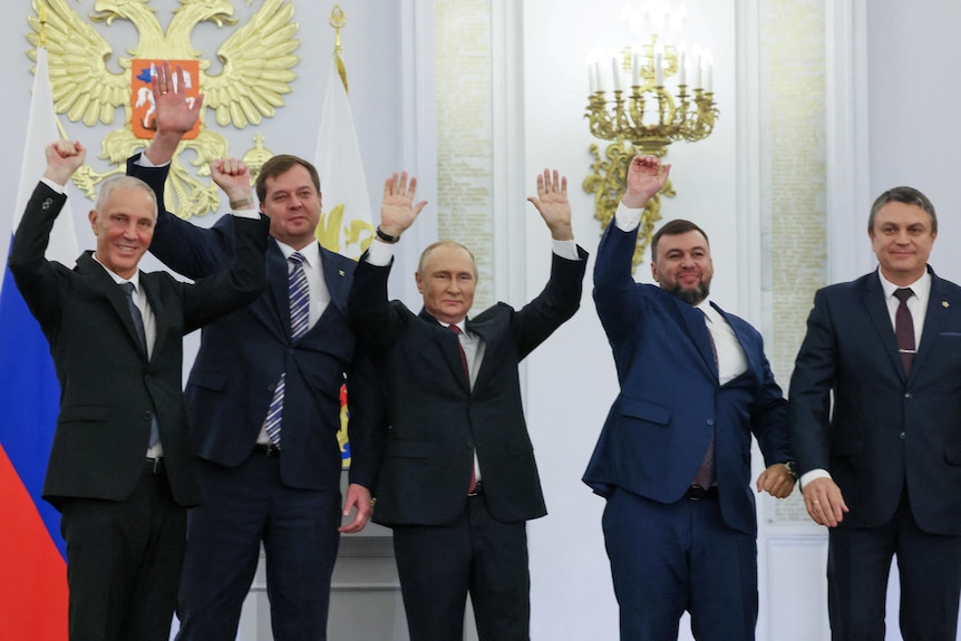 Russian president standing with four other men in suits, all with their arms up in the air.