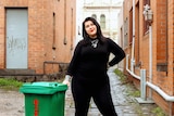 A smiling woman in black roll-neck top and trousers and lots of silver chains stands in a laneway next to a green rubbish bin. 