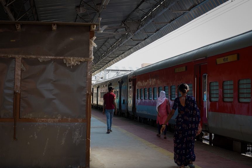 People walk past a boarded up stall on a train platform next to a red train