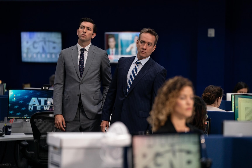 Still from TV show Succession: A man in his late 40s and a tall man in his early 30s, both in suits, stand in a newsroom