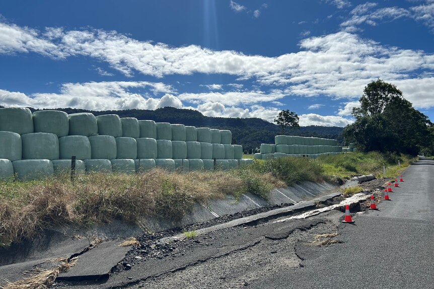 A flood-damaged bitumen road near stacks of hay bales on a farm.