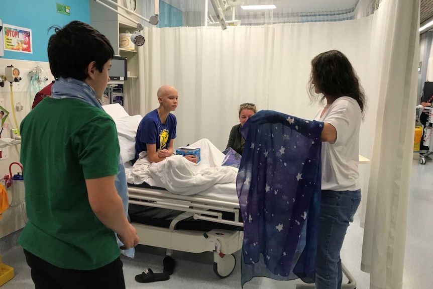 A boy and a woman with a scarf stand next to a young person in a hospital bed.