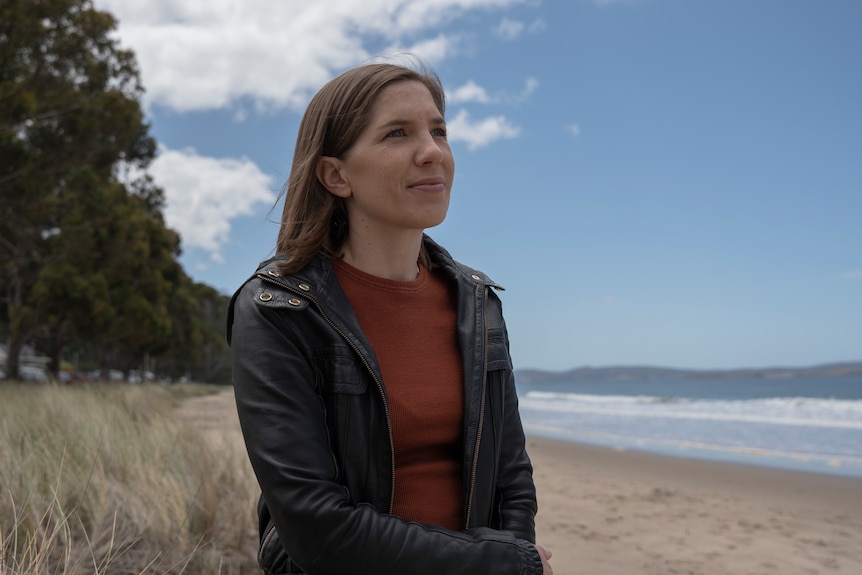 a woman in a black leather jacket on a beach on a blue-sky day, looking up with hope