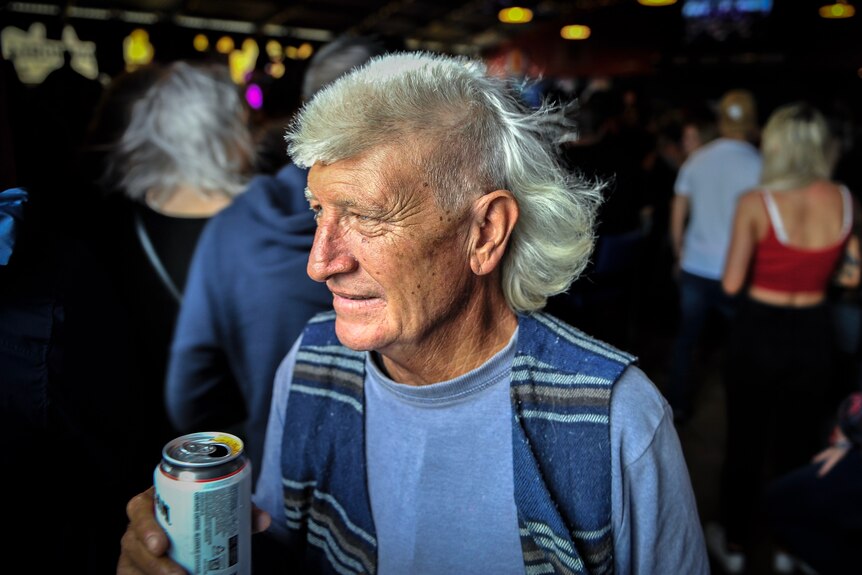 A man with grey hair and a mullet holds a can inside a pub.