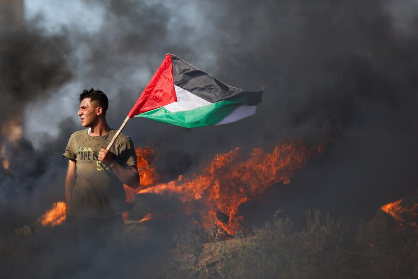 A Palestinian holds a flag in front of a fire. 