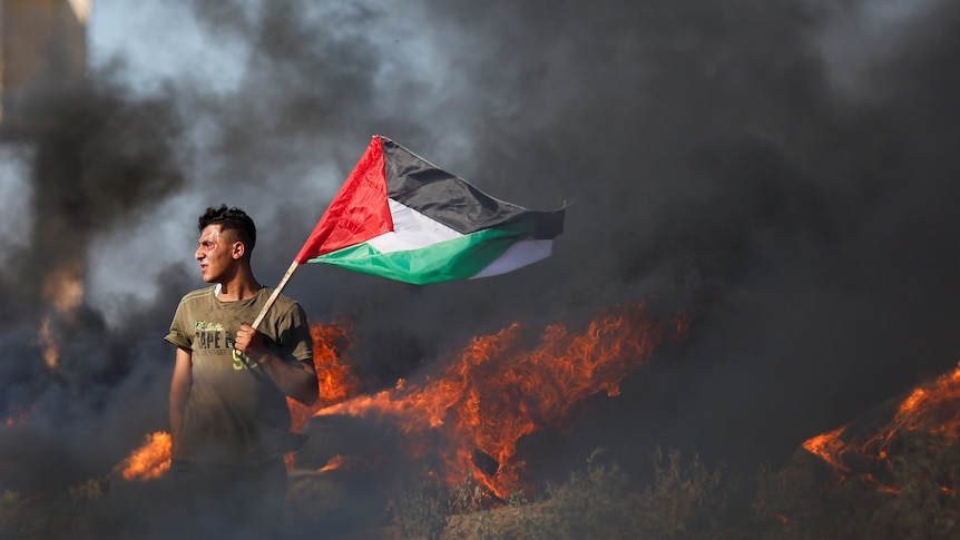 A Palestinian holds a flag in front of a fire. 