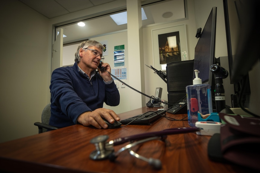 A man sits at a desk, holding a phone in one hand and using a computer with the other.