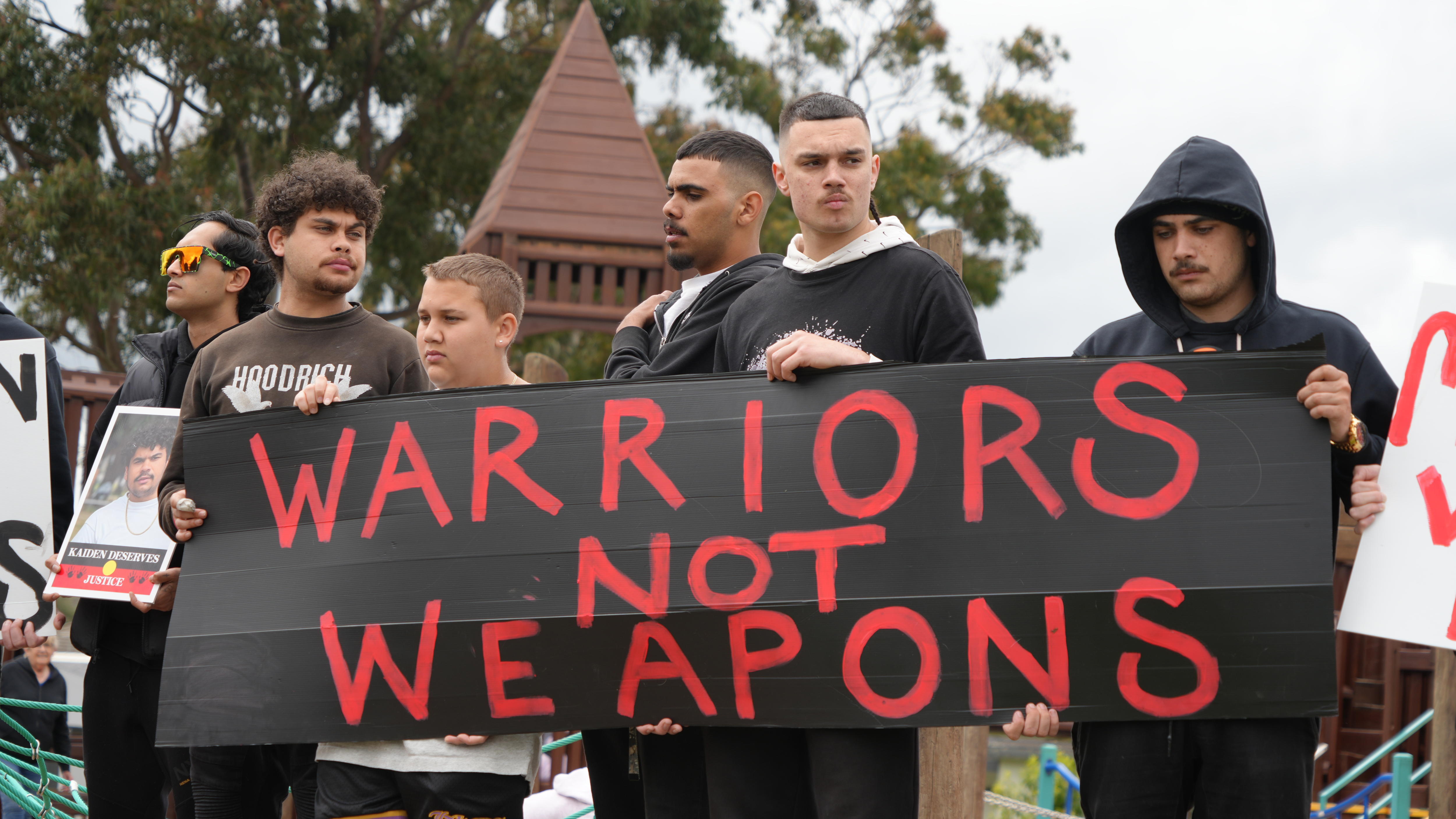 Young Aboriginal men stand with a placard reading 'Warriors not weapons'