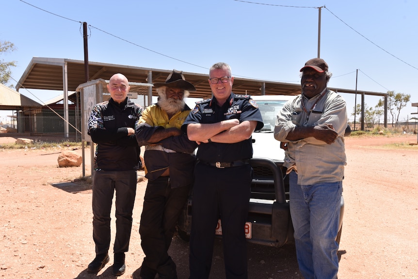 Four men smiling standing in front of car parked on red dirt in remote central australian town