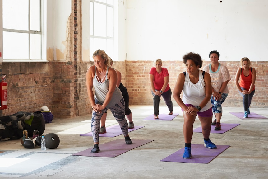 A group of women in a gym class stand on mats, and are doing lunges.