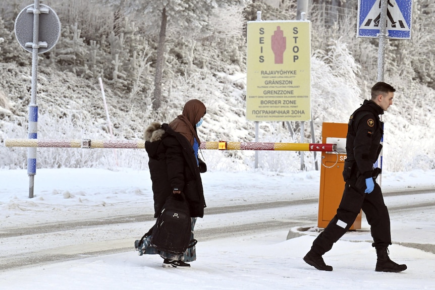 A woman carrying a backpack walks behind a border security guard on a snowy road