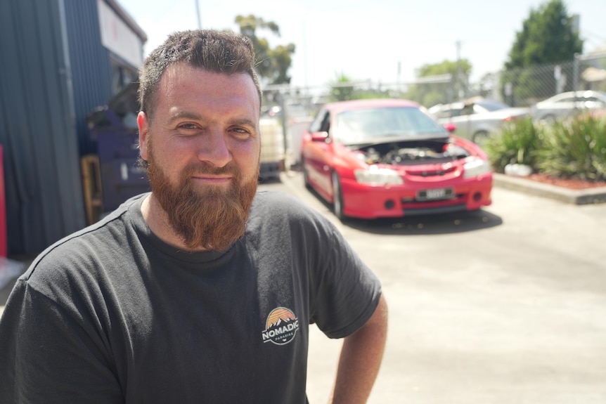 Jayden Clark wearing a grey shirt, sporting a beard and pictured in front of a car mid-repairs