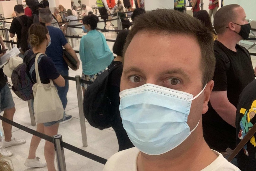 A man in a facemask takes a selfie in front of a queue of passengers in an airport terminal.