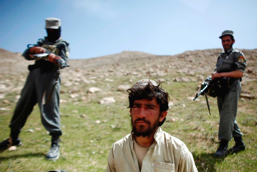 A bearded man in a white shirt stands in front of two soldiers in point guns at him in a field.