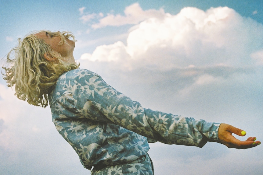 A 2022 press shot of HAAi with arms outstretched to the right against a cloudy blue sky