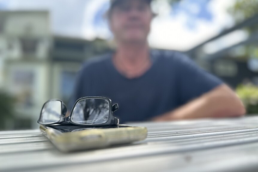 A close up of a pair of glasses and a phone with a man out of focus in the background. 