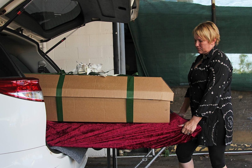 A photo of Carol Phayer as she loads a cardboard coffin into her a converted hearse.